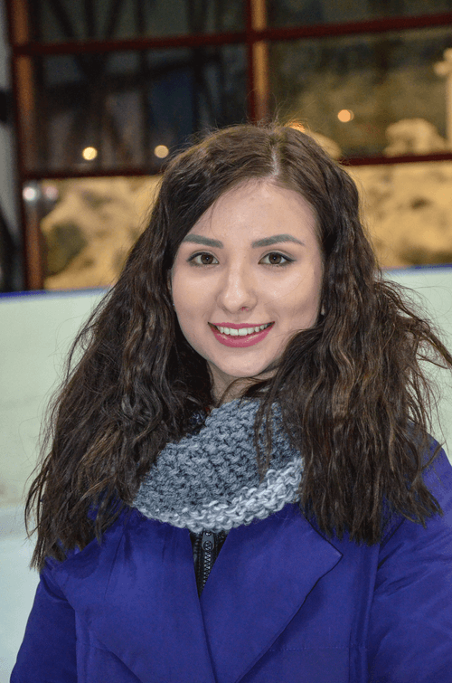Smiling woman with long curly dark hair wearing a blue coat and gray knitted scarf.
