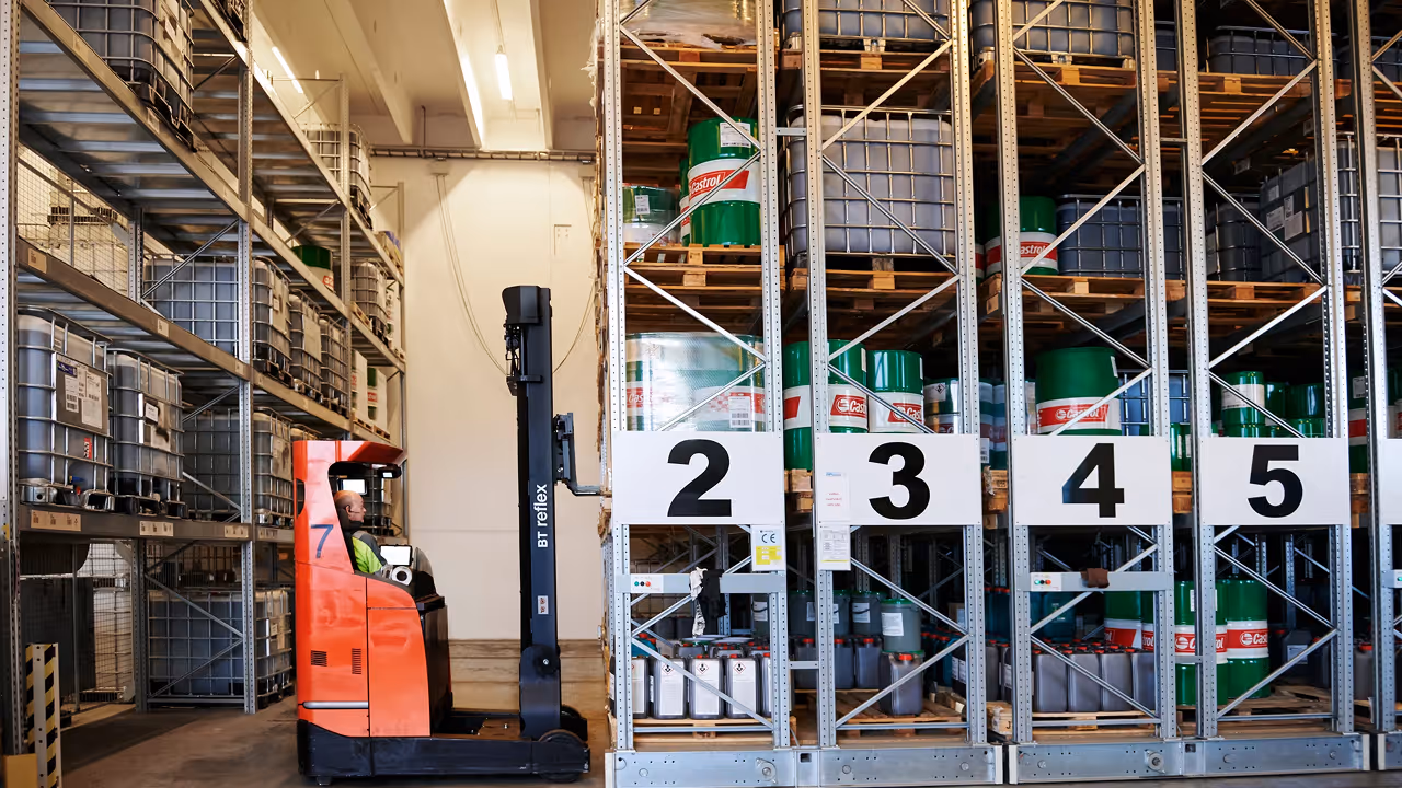 Vīrietis vadā kravas pacēlāju noliktavā pie plauktiem ar zaļām Castrol mucām.