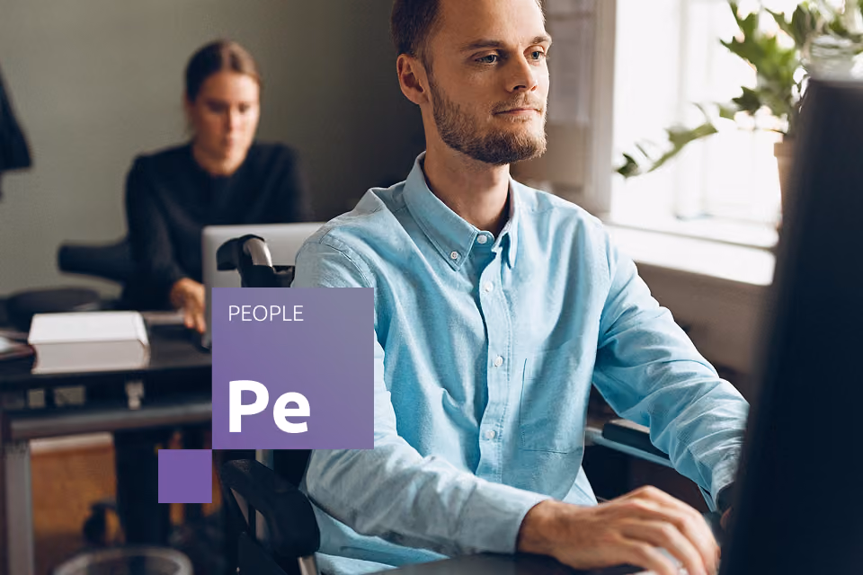 Cilvēks ratiņkrēslā strādā pie datora birojā, fonā vēl viens darbinieks.