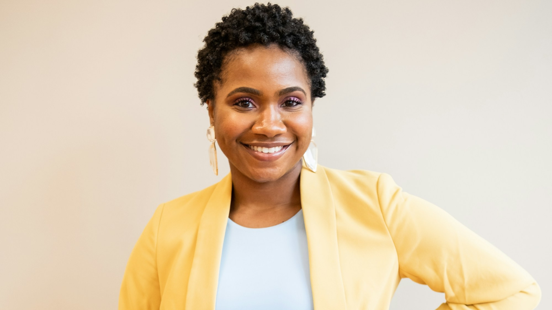 Smiling woman with short curly hair wearing a yellow blazer and light blue top against a neutral background.