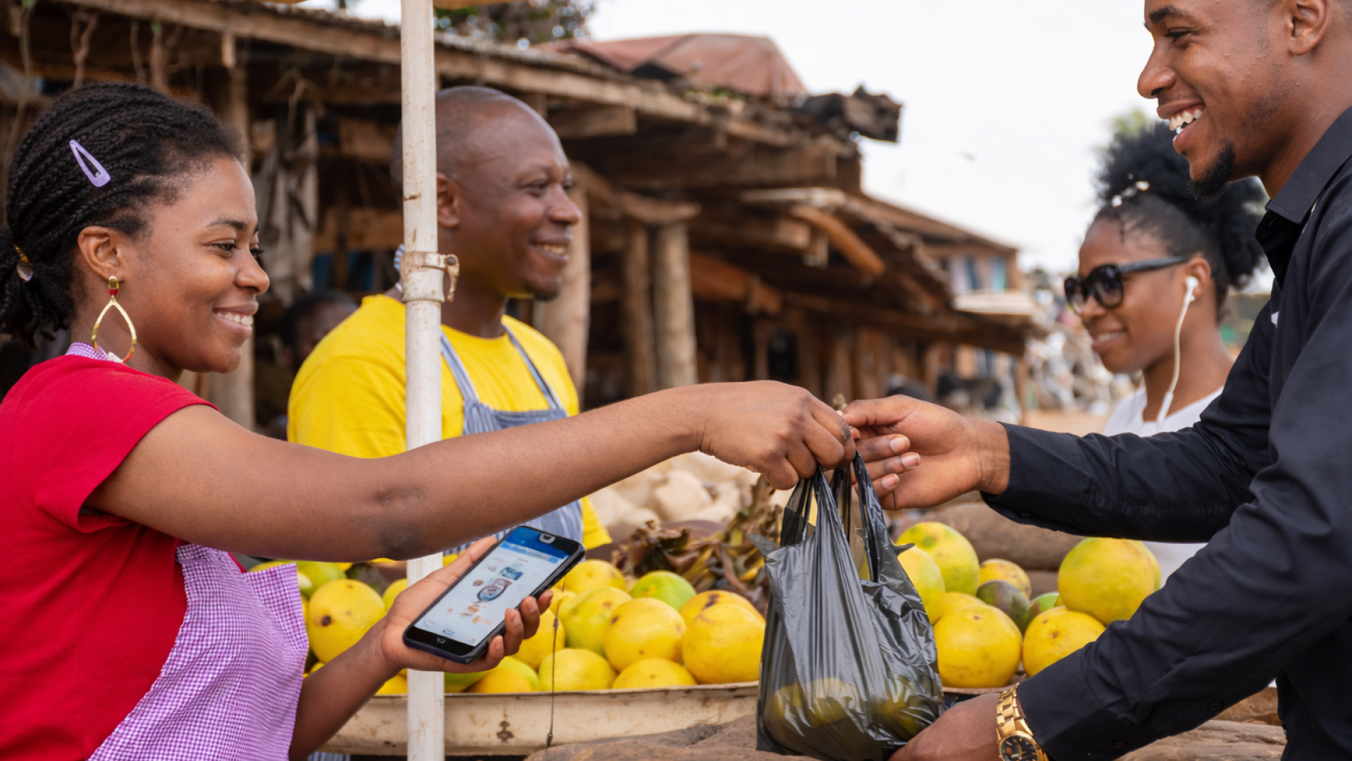 A woman vendor handing a black plastic bag to a smiling man at an outdoor market with yellow fruits in the background.
