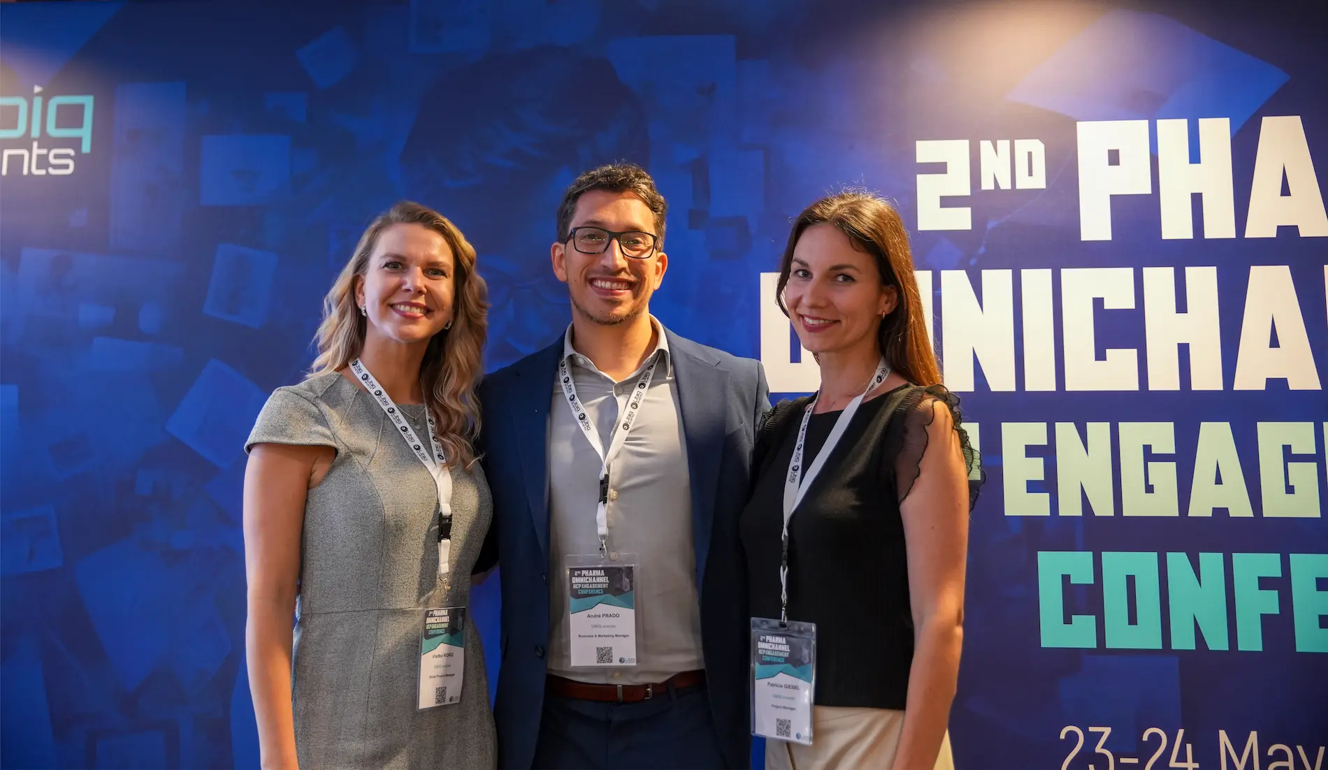Three smiling conference attendees wearing name badges standing in front of a blue backdrop with '2nd Pharma Omnichannel Engagement Conference' text.