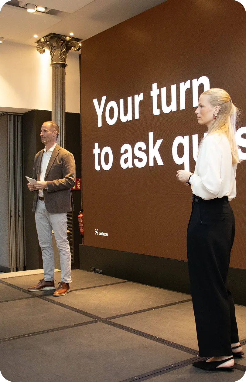 Two presenters on stage with a large screen behind them displaying the text 'Your turn to ask questions.'