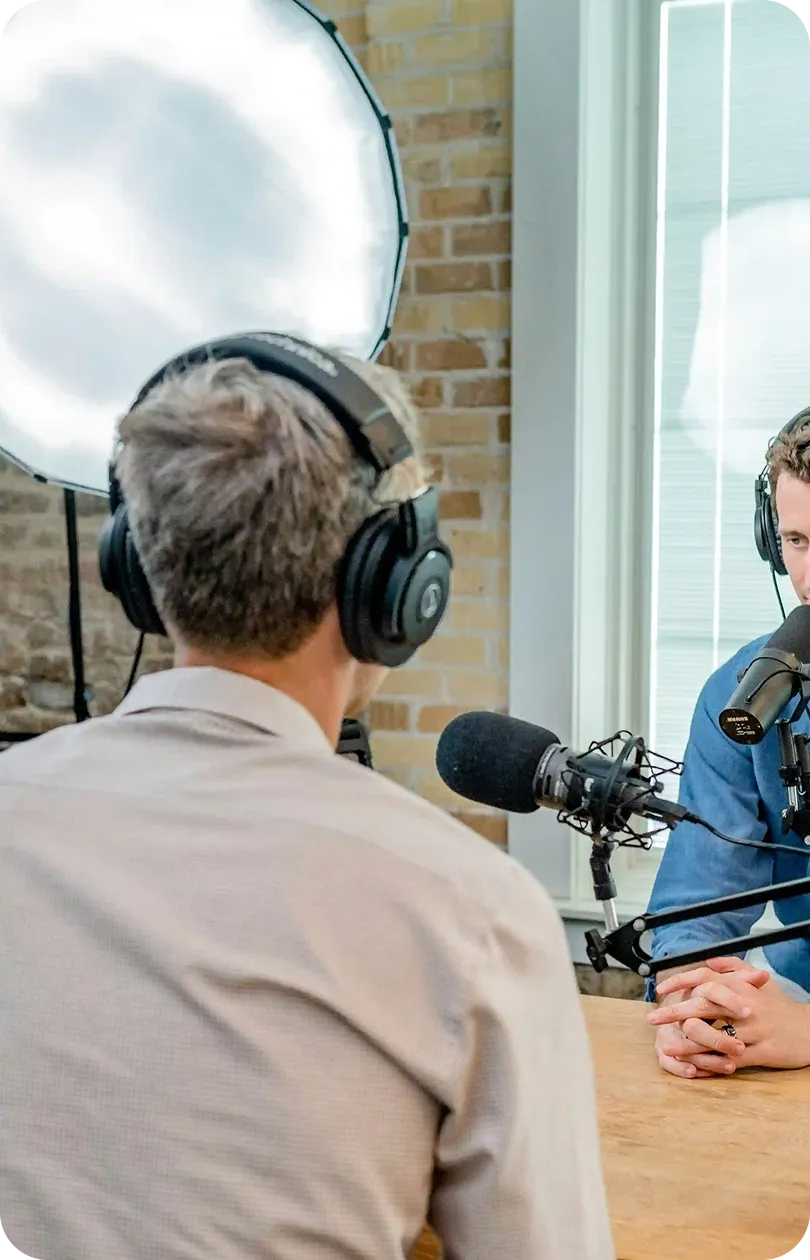 Two people wearing headphones sitting across from each other at a table, speaking into microphones in a podcast or recording studio setup.