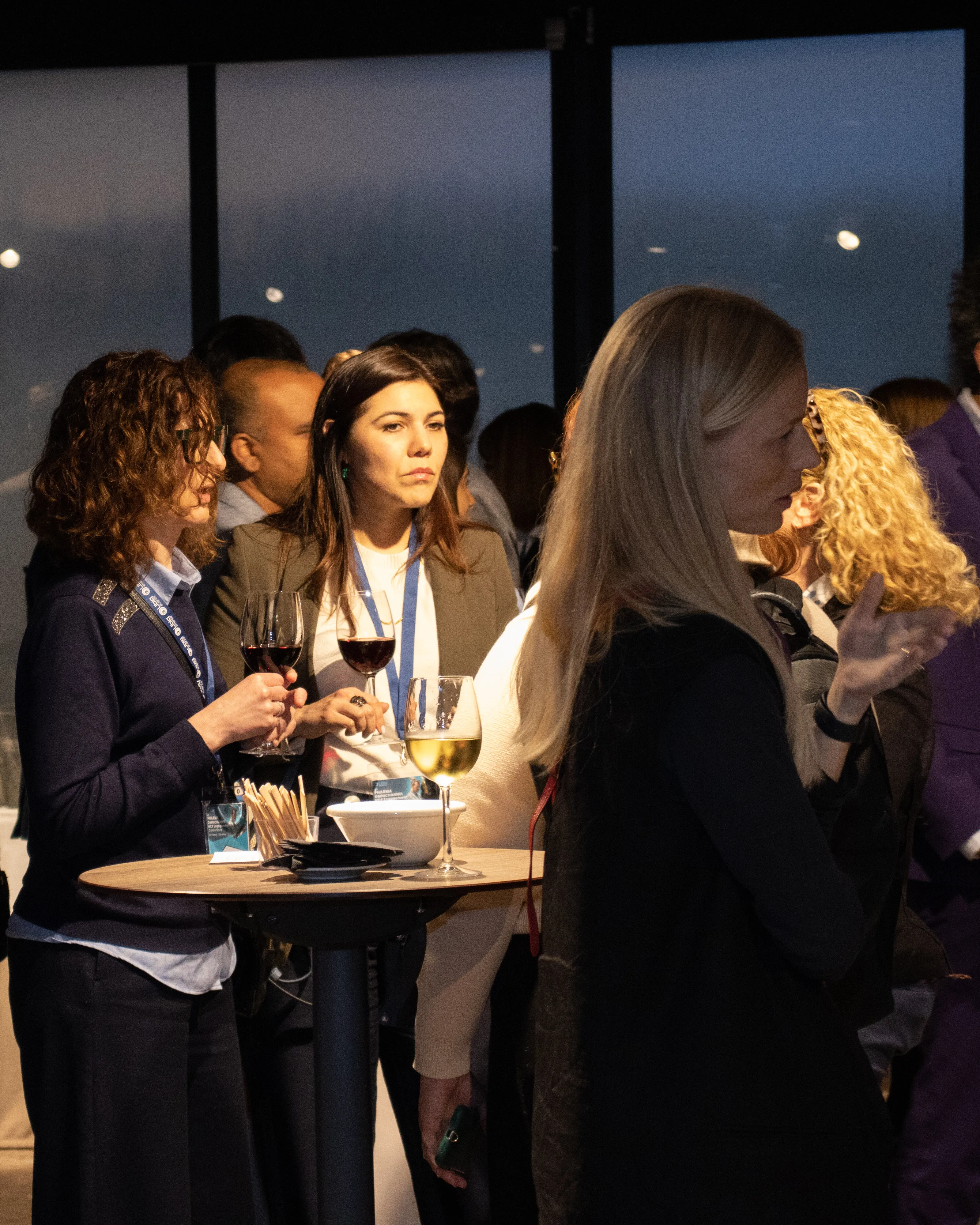 People socializing at a cocktail event holding glasses of red and white wine around a high table with snacks.