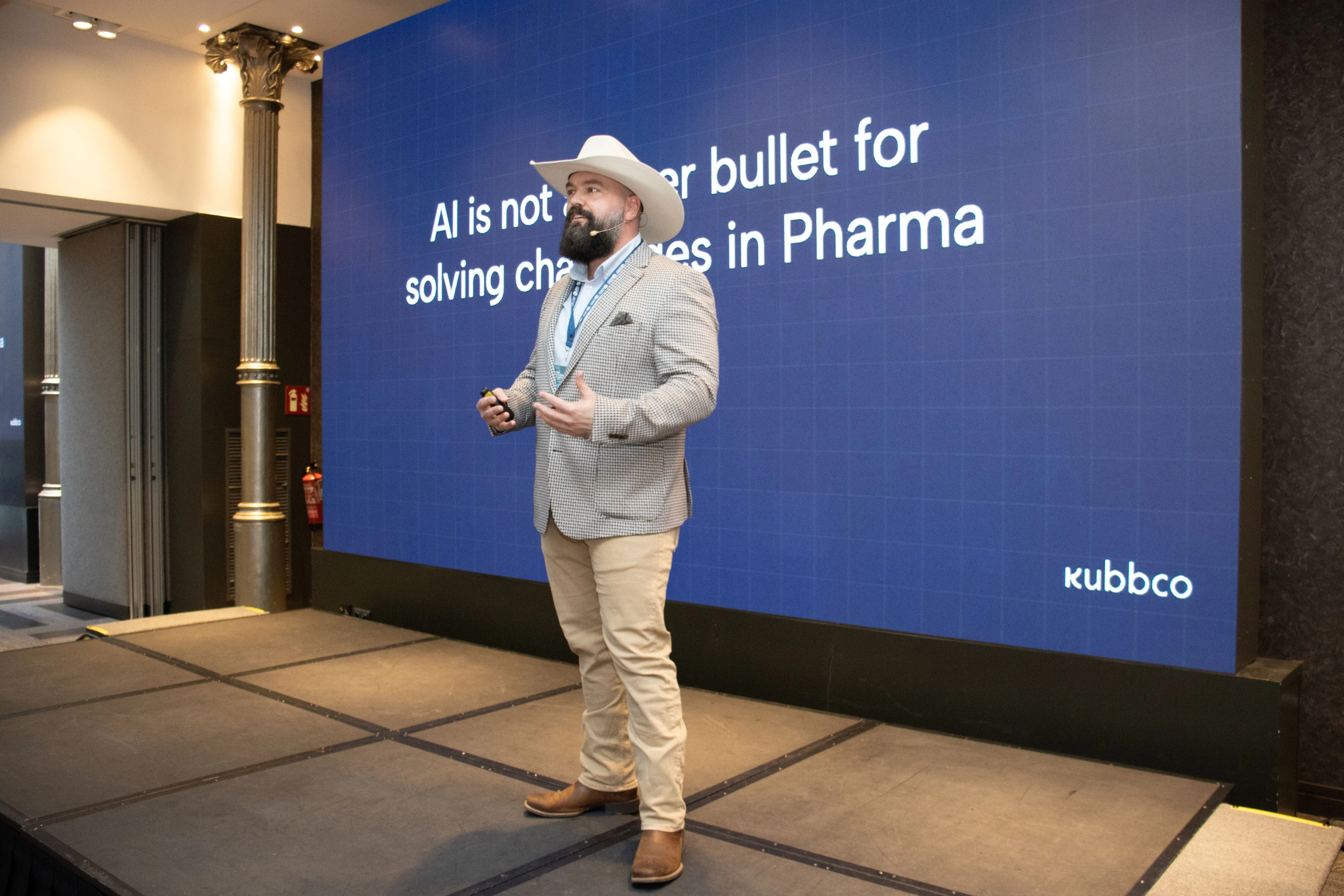 Man wearing a cowboy hat and checked blazer speaking on stage in front of a large screen that reads 'AI is not a silver bullet for solving challenges in Pharma'.