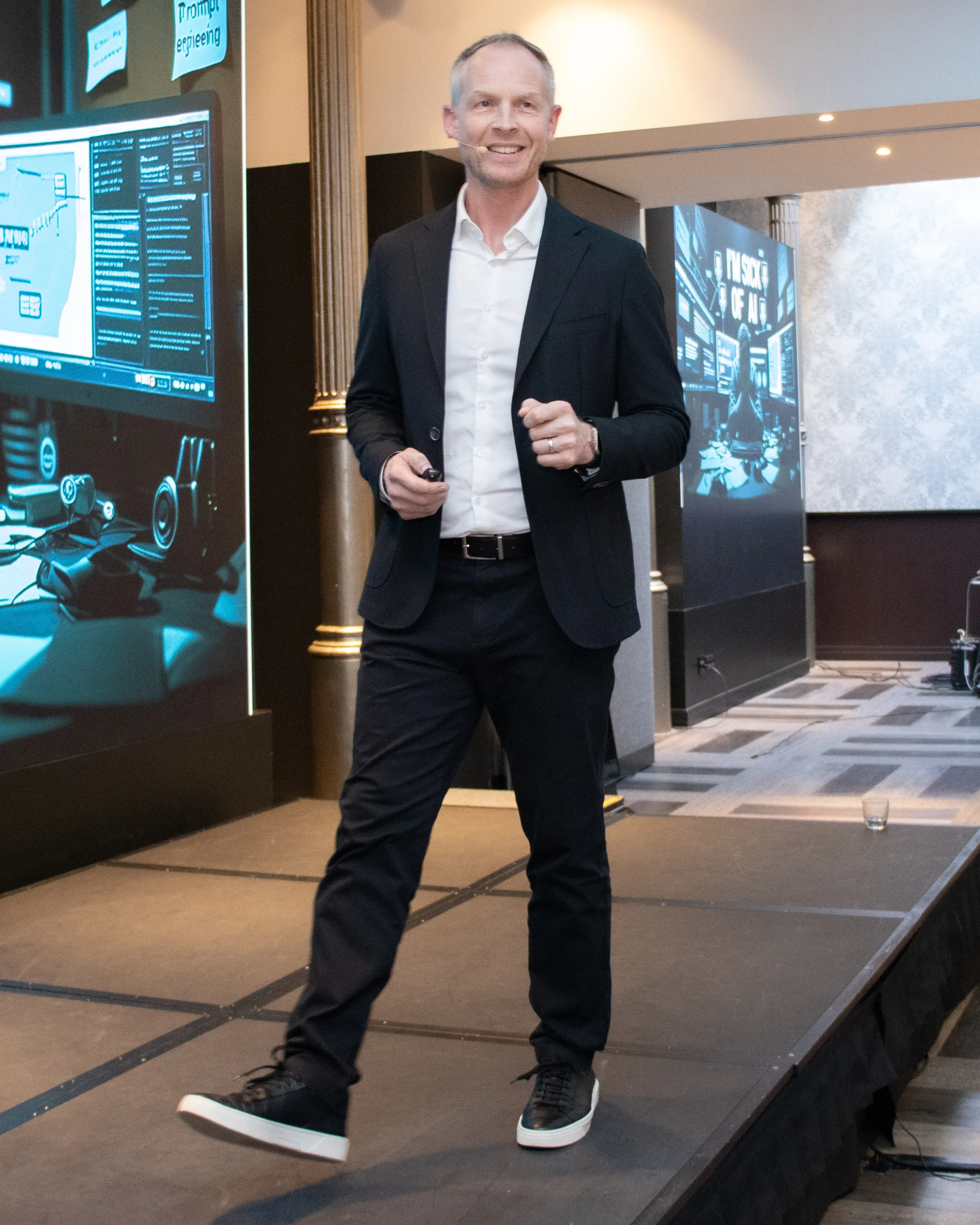 Smiling man in a black suit and white shirt walking on a stage with a microphone headset and presentation screens in the background.