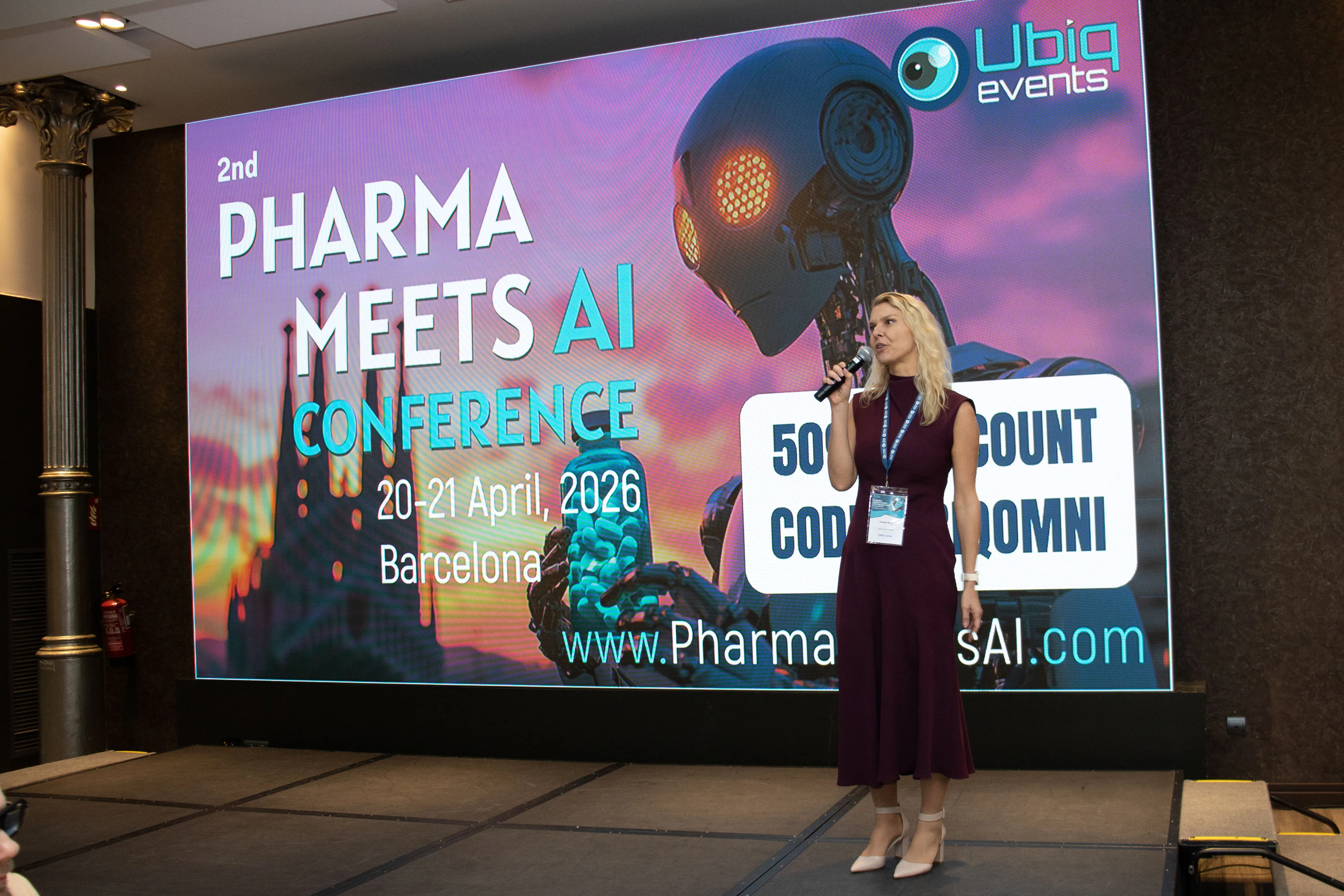 Woman with microphone speaking on stage in front of a large screen displaying '2nd Pharma Meets AI Conference, 20-21 April 2026, Barcelona' with a robot graphic and Ubiq events logo.