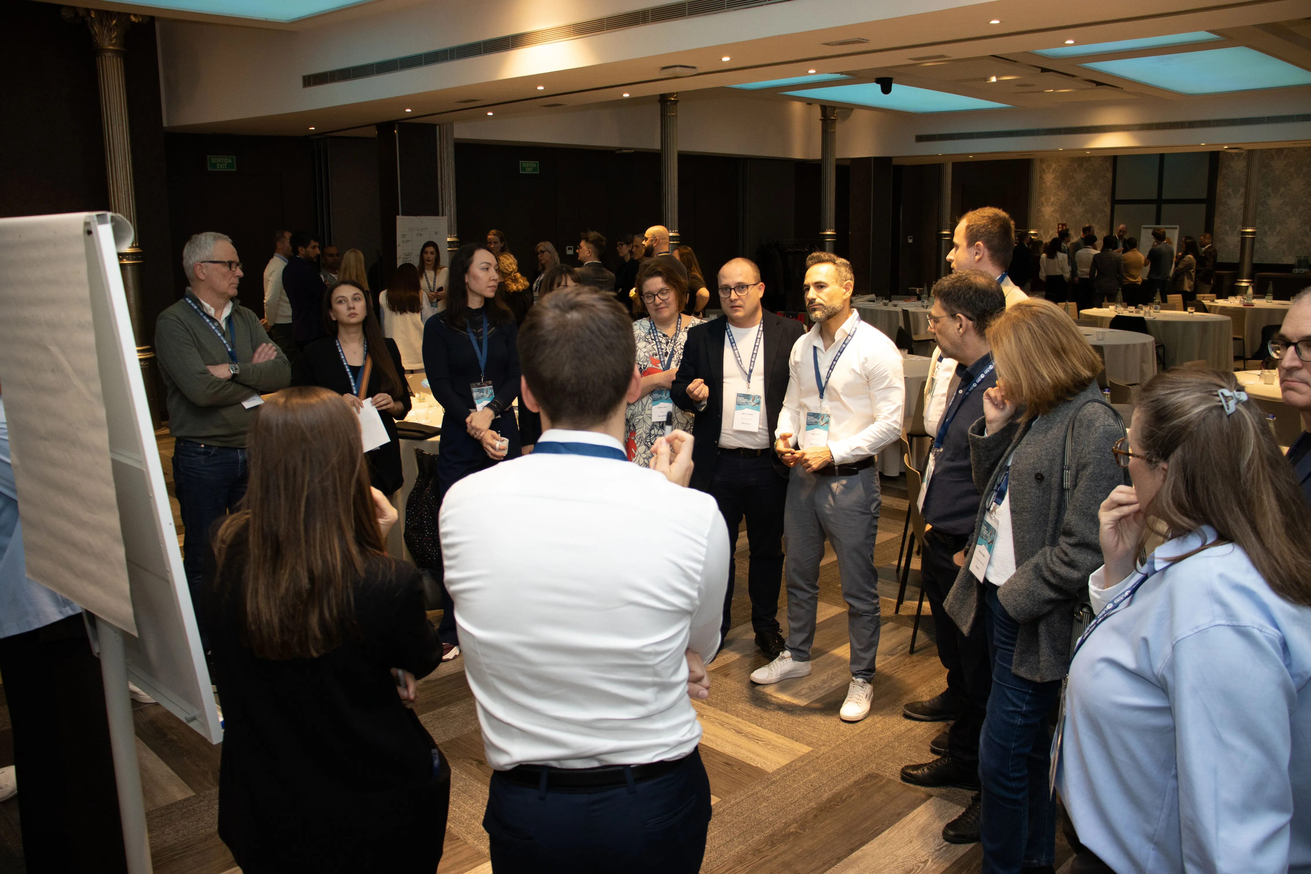 Group of professionals in business attire standing in a conference room, engaged in discussion near a blank flip chart.