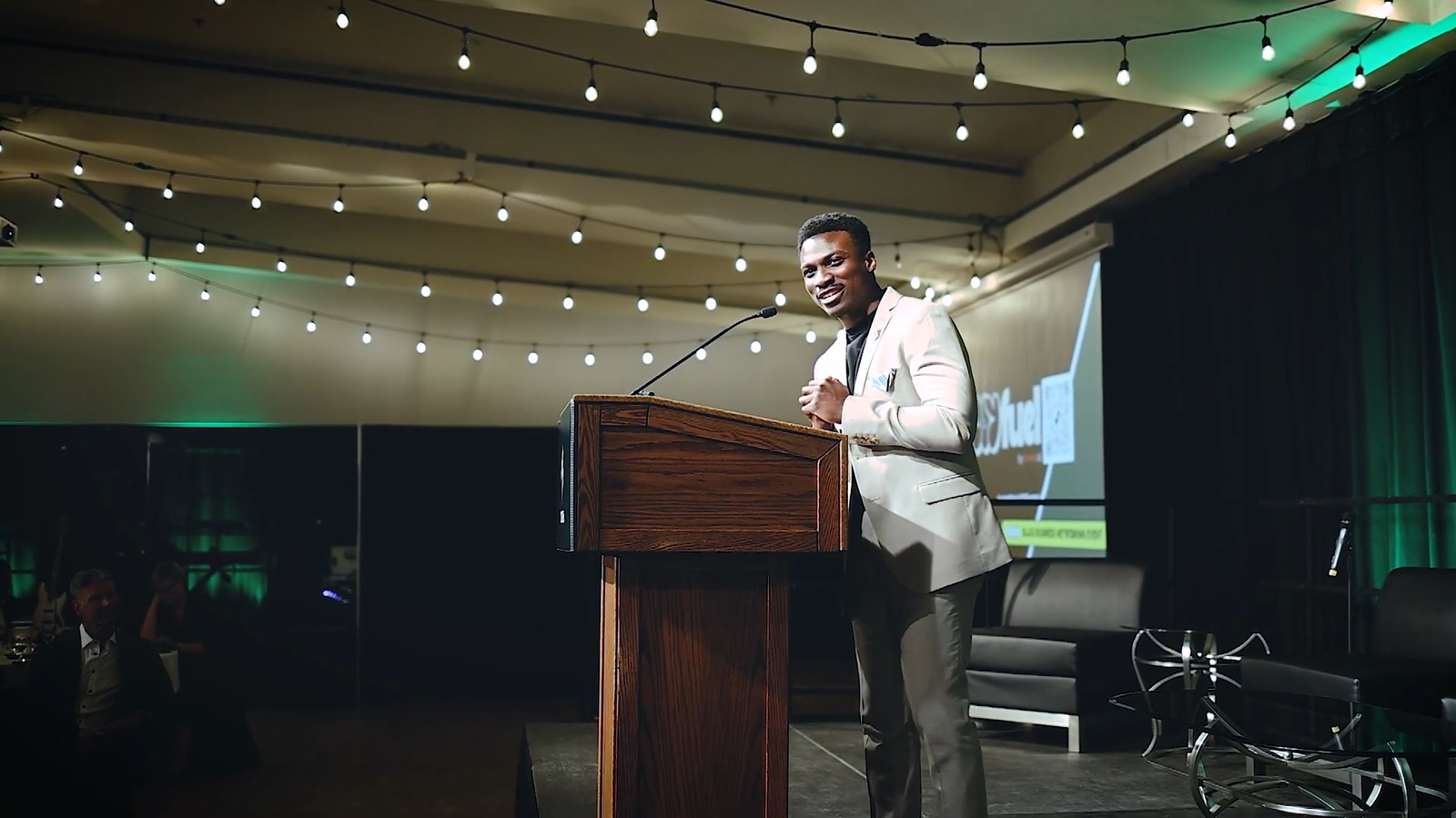 David Akinmade smiling at the audience from a lectern under string lights, the JOYfuel logo on the screen behind him
