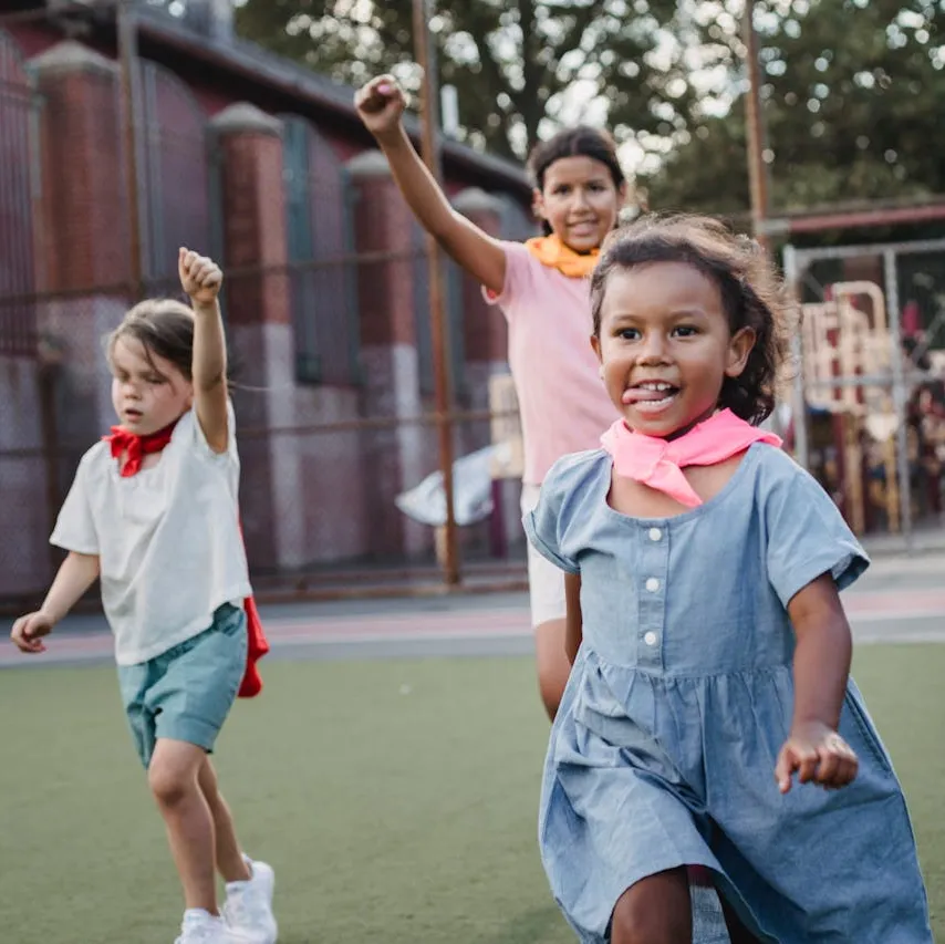 Three children playing outdoors on a grassy field, smiling and raising their fists, with colorful bandanas around their necks.
