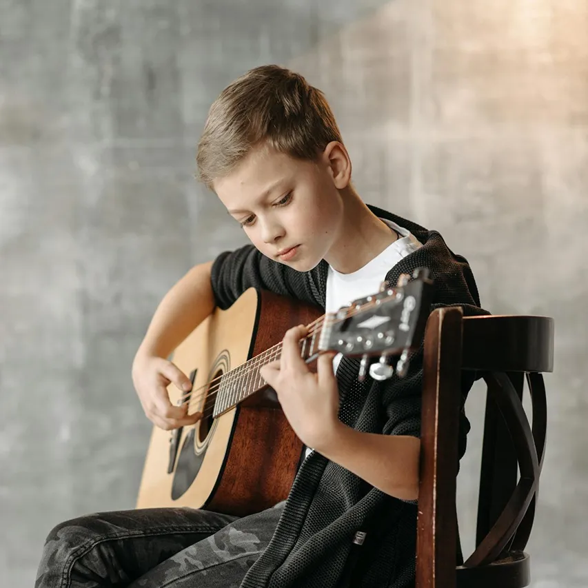 Young boy sitting on a wooden chair playing an acoustic guitar with focused expression.