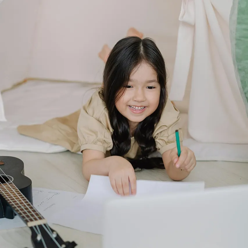 Smiling young girl lying on the floor drawing on paper with a green pencil, with a guitar nearby.