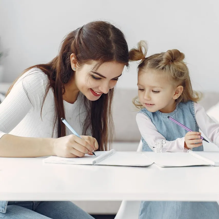 Young woman and little girl sitting at a white table, writing in notebooks together with colored pencils.