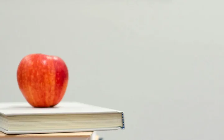 Side view of an open laptop leaning against a stack of three thick books on a wooden surface with a white brick wall background.