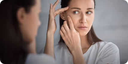 Kobieta w białej koszulce ogląda twarz w lustrze, dotykając policzka i czoła.