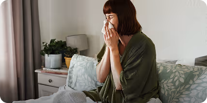 Kobieta osłaniająca nos chusteczką siedzi na kanapie w domu, wyglądając na przeziębioną.