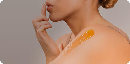 Kobieta nanosząca przezroczysty żel na ramię palcem, w tle jasne neutralne tło.