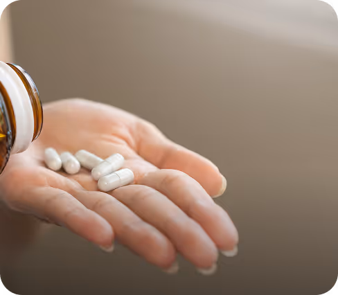 Ręka osoby trzymająca pięć białych kapsułek wyciągniętych z brązowej butelki.