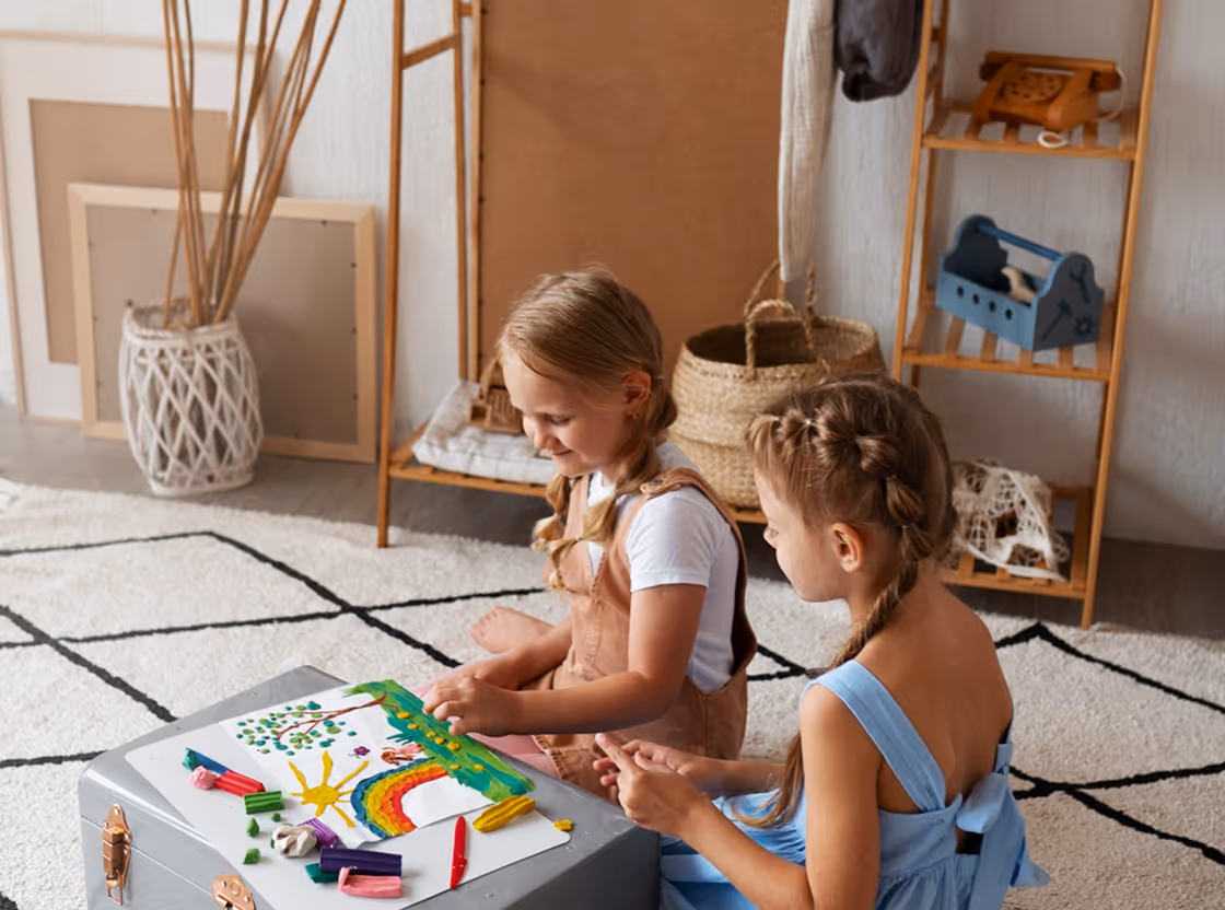 Two young girls sitting on a rug, drawing and coloring a rainbow and sunny landscape on paper with crayons on a small gray table.