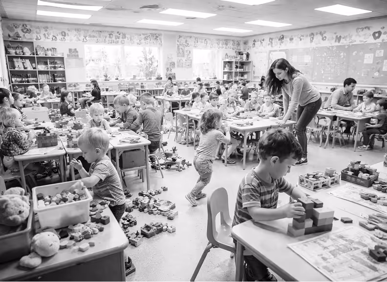 Classroom full of young children seated at tables playing with building blocks while a teacher assists them.
