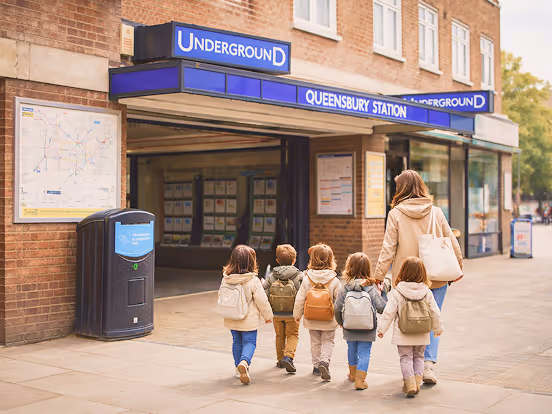 Group of children with backpacks and an adult walking towards Queensbury Station entrance of the London Underground.