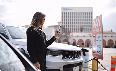 Woman using phone to pay at a parking meter beside white SUV on city street.