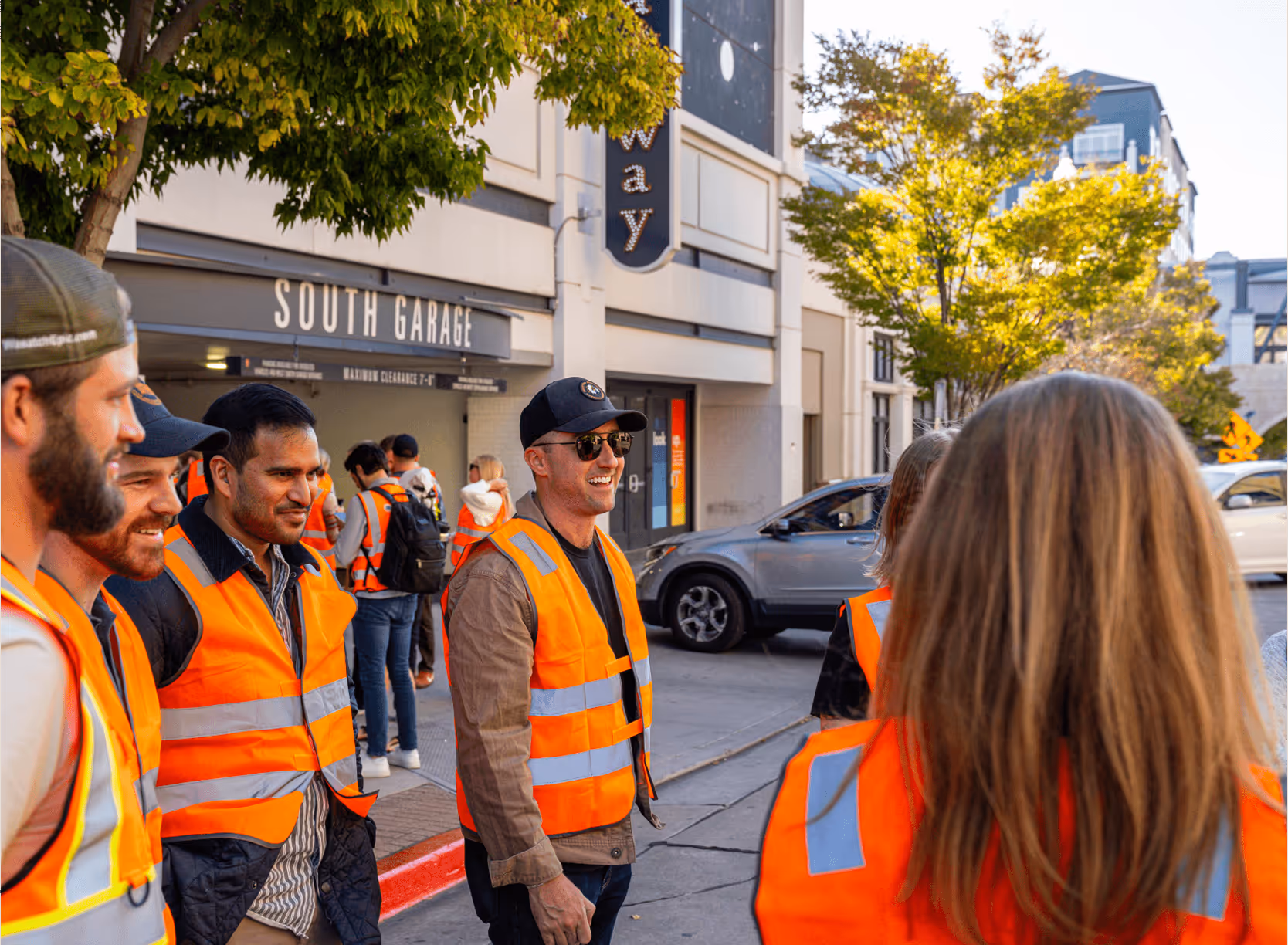 Group of people wearing bright orange safety vests standing and talking near a parking garage entrance.