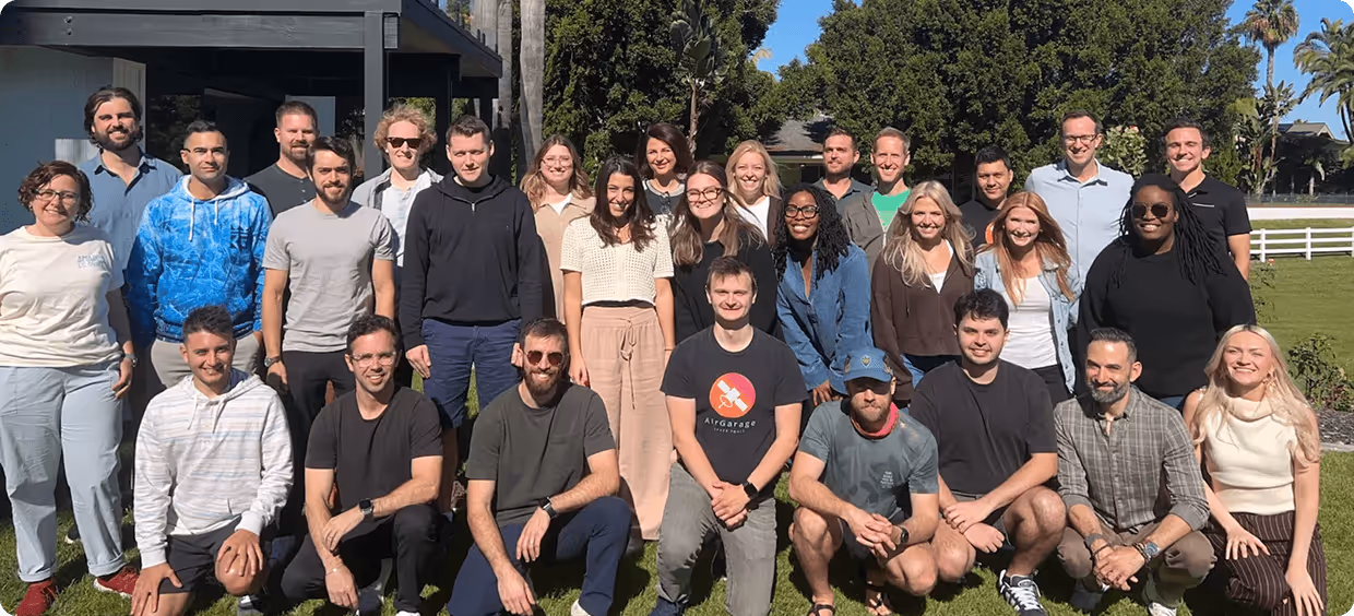 Group of diverse people posing outdoors on grass with trees and a white fence in the background.