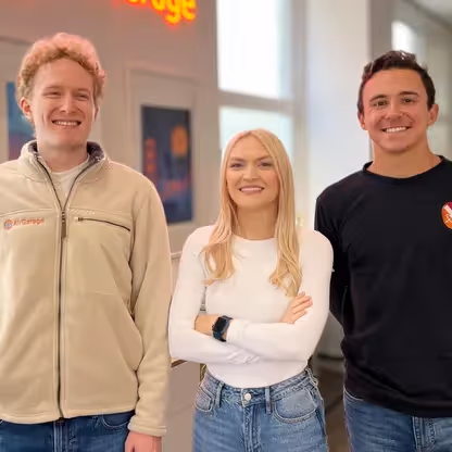 Three smiling young adults standing indoors, two men on sides and a woman in the middle with arms crossed.