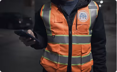 Person in an orange reflective safety vest holding a smartphone in a dimly lit parking area.