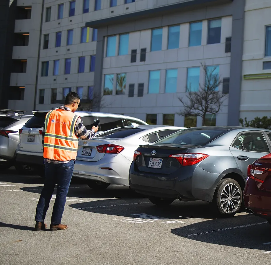Man in orange safety vest inspecting parked cars in a parking lot next to a building.