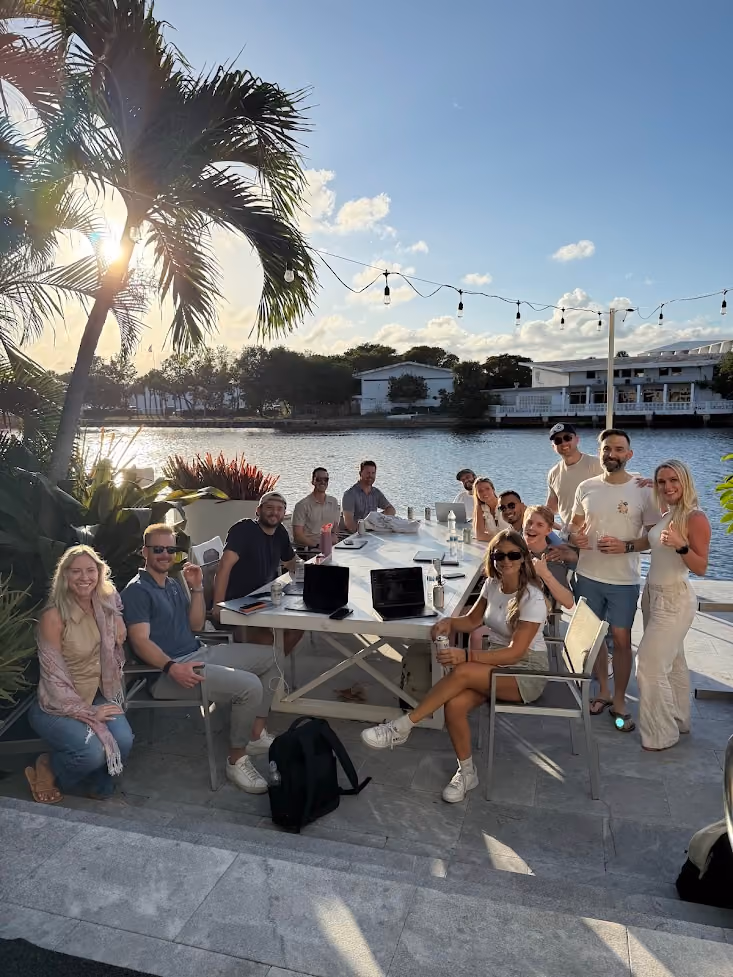 Group of people sitting and standing around a table outdoors by water with laptops and drinks at sunset.