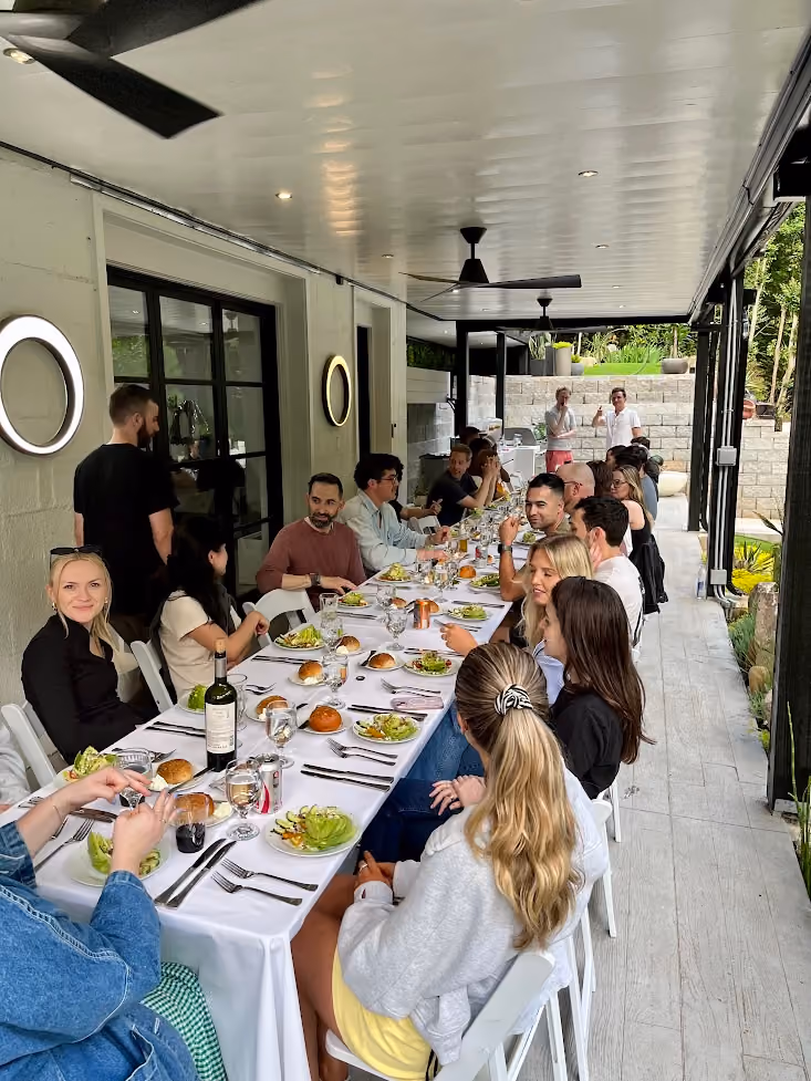 Group of people seated at a long outdoor table set with salad plates and drinks under a covered patio.