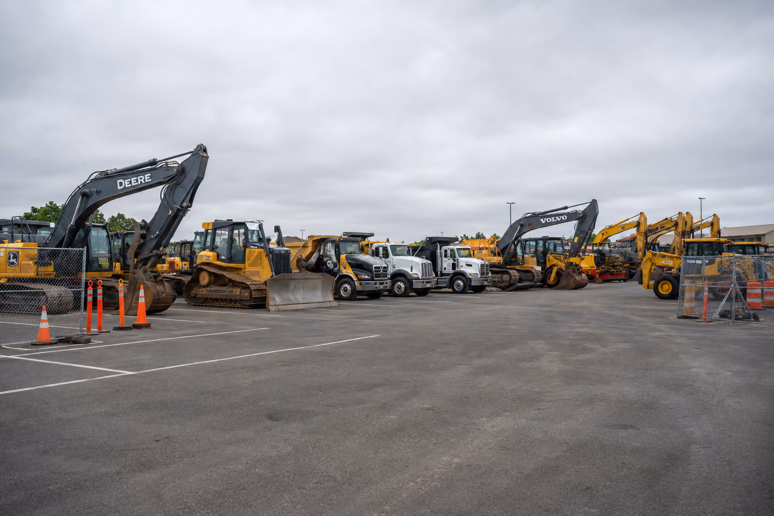 Heavy construction equipment and trucks lined up in a parking lot under a cloudy sky.