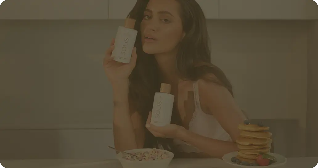 Woman in white top holding two SOLIS bottles sitting at a kitchen table with pancakes and cereal.