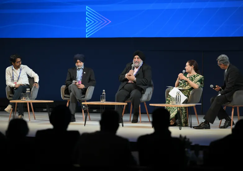 Five people seated on stage panel discussion, speaking with microphones, audience silhouettes in foreground.