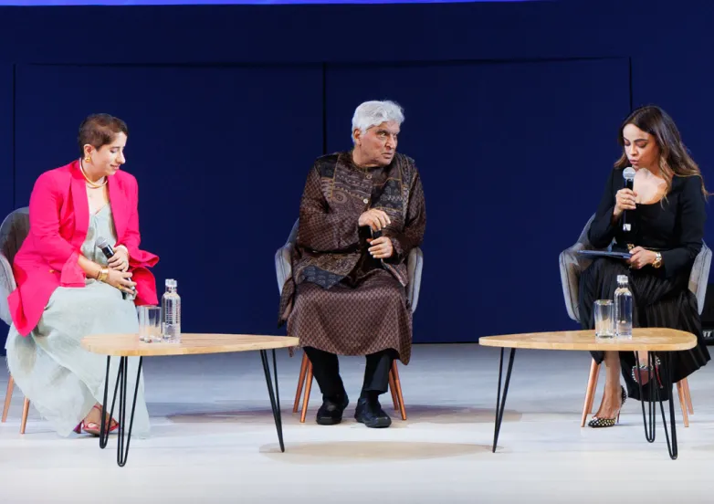 Three people seated on stage chairs having a discussion, each holding a microphone, with two small tables in front.