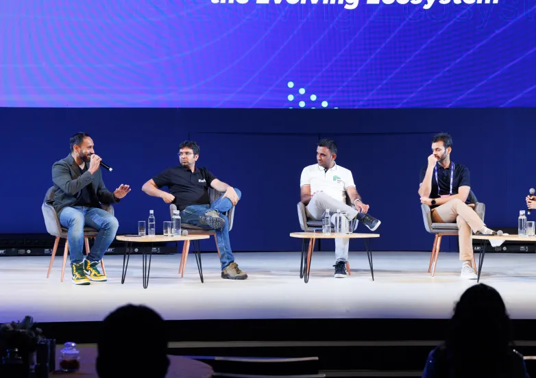 Four men seated on stage having a panel discussion at a conference with microphones and water bottles.