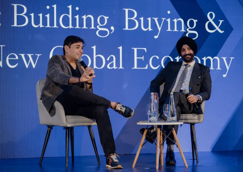 Two men seated on stage speaking into microphones at a business event with blue backdrop text.