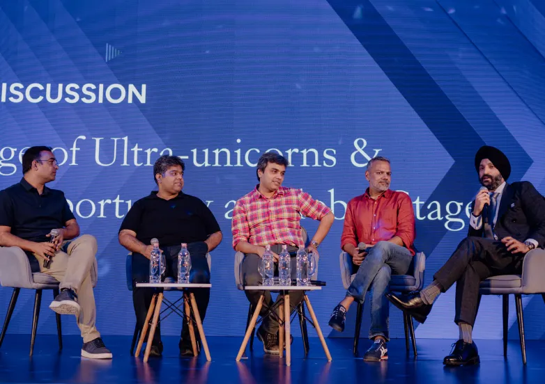 Five men seated on stage in a panel discussion, holding microphones, with water bottles on tables.