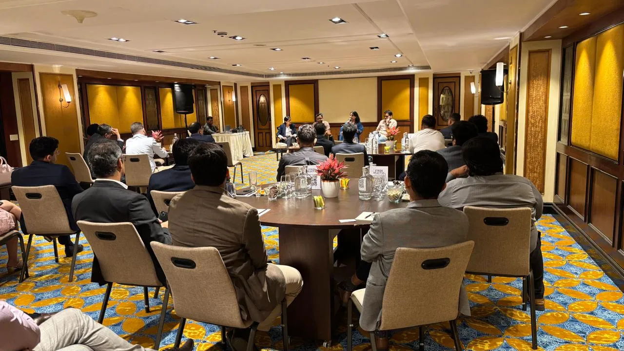 People seated around tables listening to a panel discussion in a carpeted conference room.