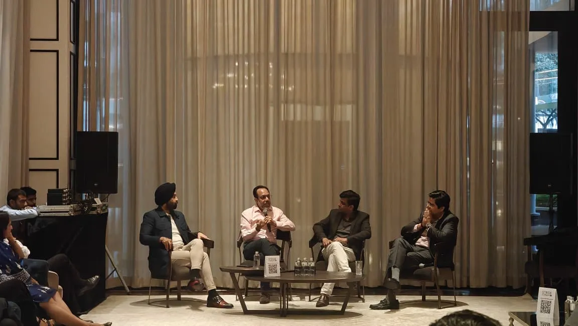Four men seated on chairs in a panel discussion in front of beige curtains in a conference room.