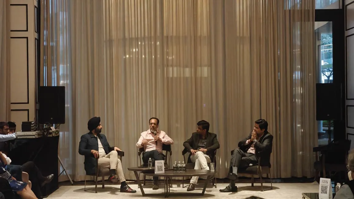 Four men seated on chairs in a panel discussion with a table and water bottles in front of them.