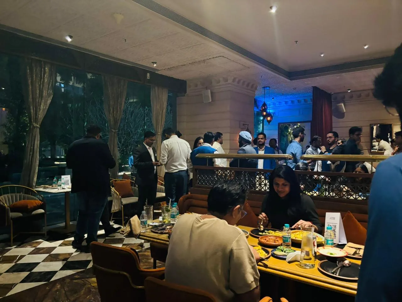 People dining and socializing in a warmly lit restaurant with modern decor and large windows.