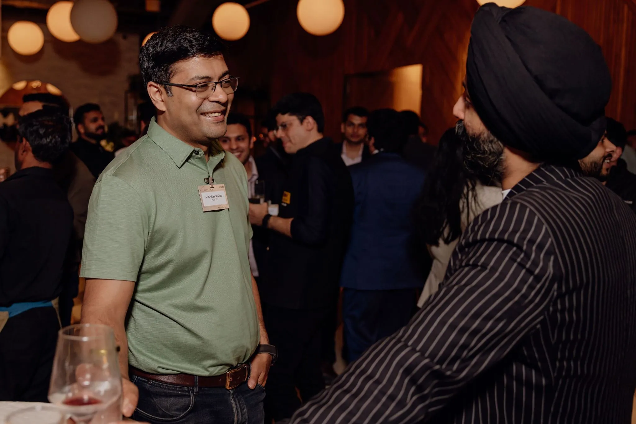 Two men smiling and talking at a social event with others mingling in a warmly lit room.