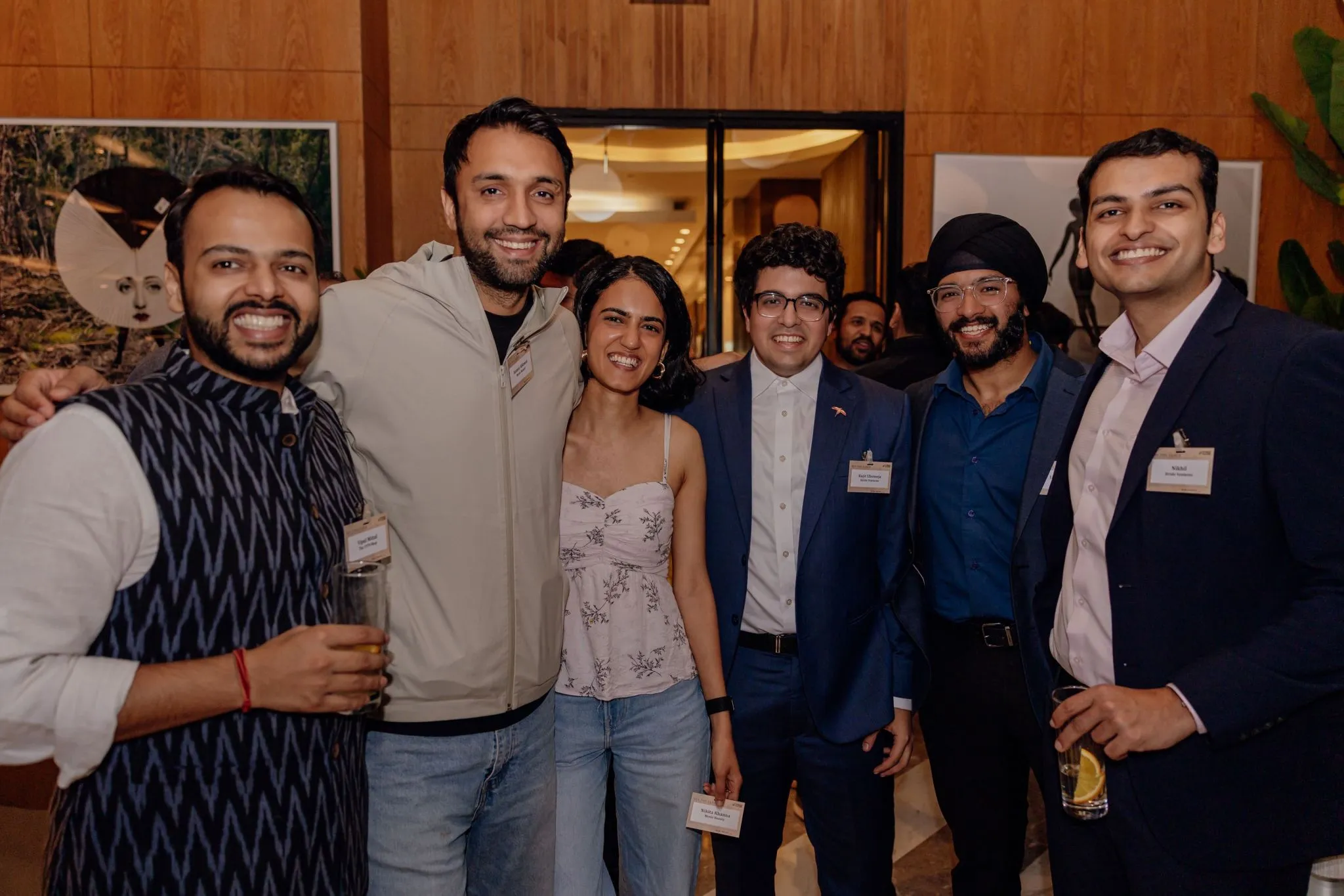 Six smiling people posing closely at an indoor event, some holding drinks, wearing name tags.