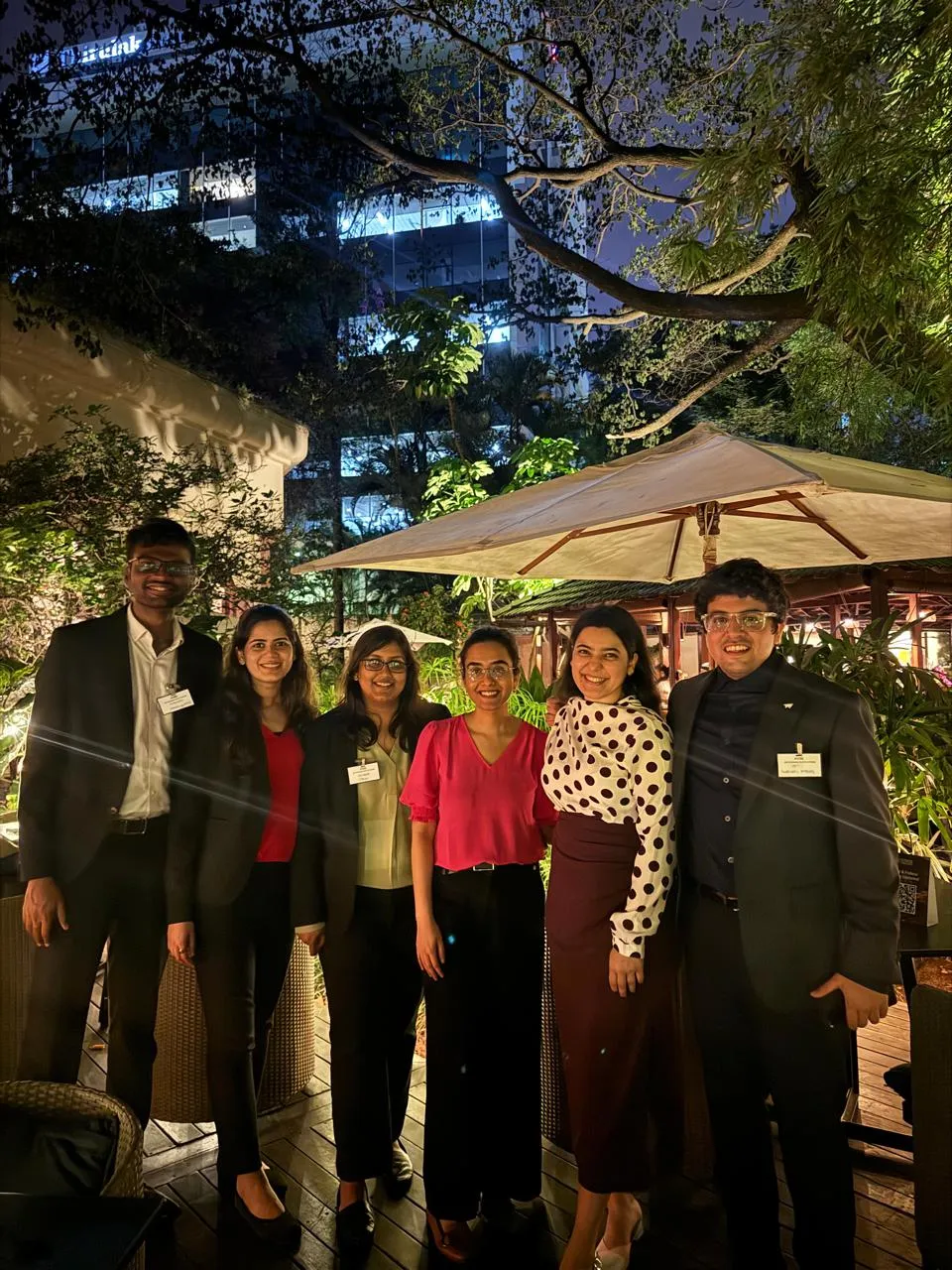 Group of six adults dressed formally, standing outdoors at night with greenery and umbrellas behind them.