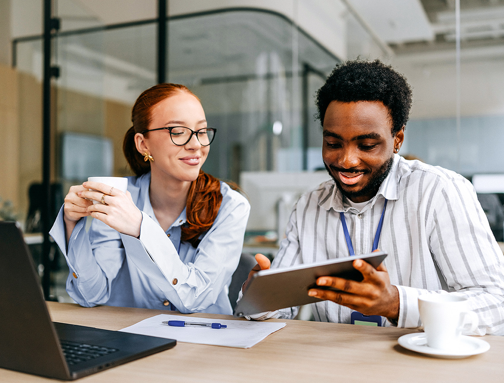 Zwei Kollegen sitzen an einem Schreibtisch, einer hält eine Tasse und der andere zeigt etwas auf einem Tablet.
