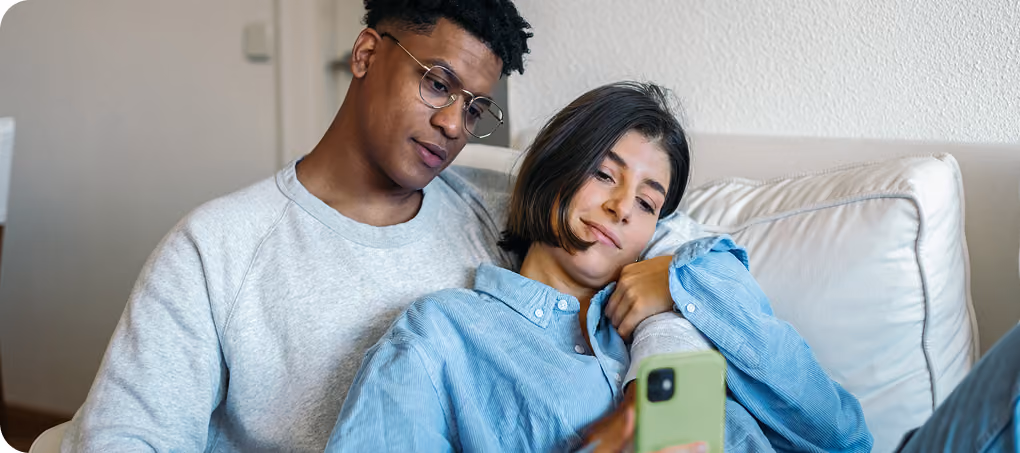 Ein Paar sitzt gemütlich auf einem Sofa, die Frau lehnt sich an den Mann und beide schauen auf ein Smartphone.